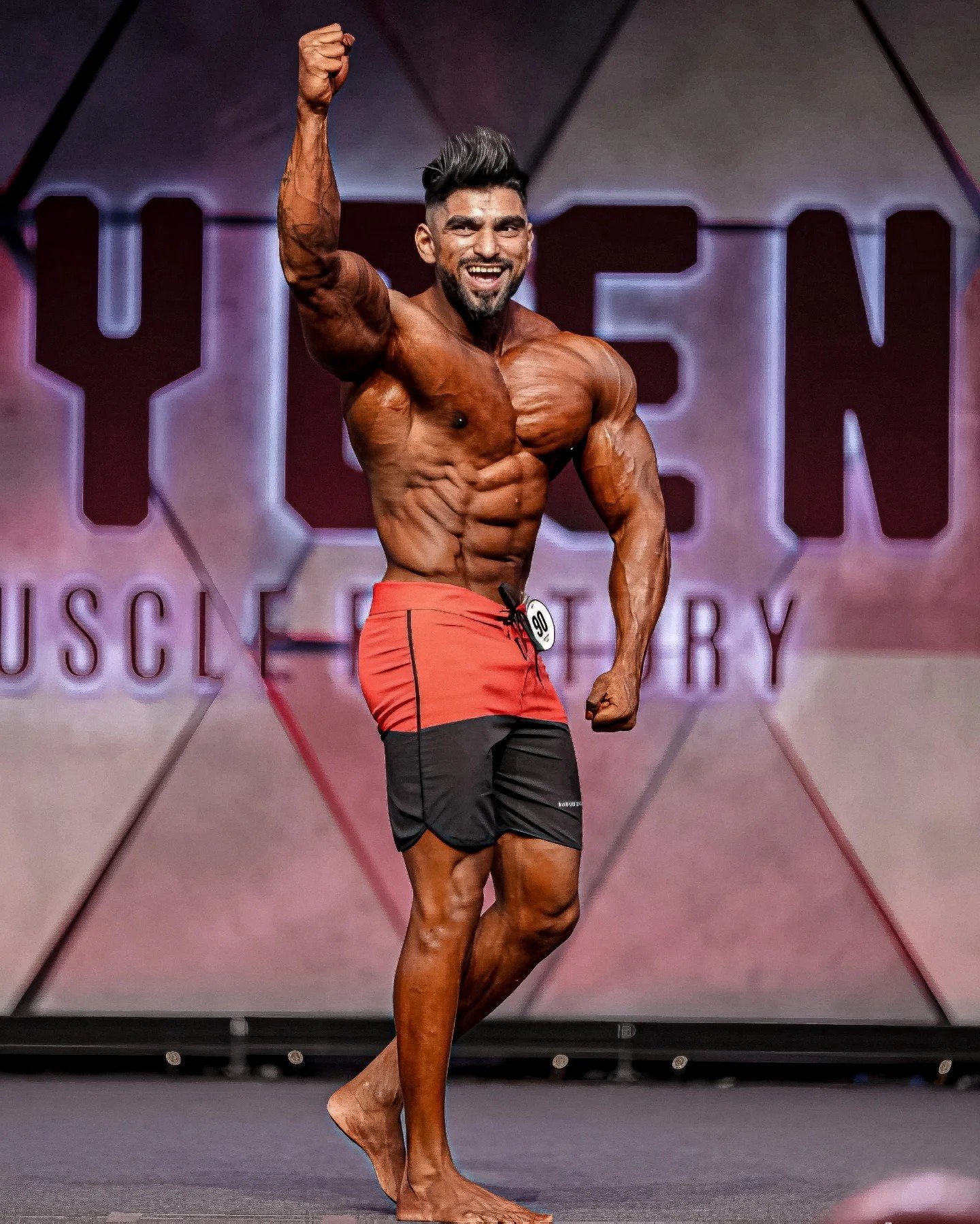Amit Agre celebrating on stage in red and black board shorts, fist raised, wearing competitor number 90