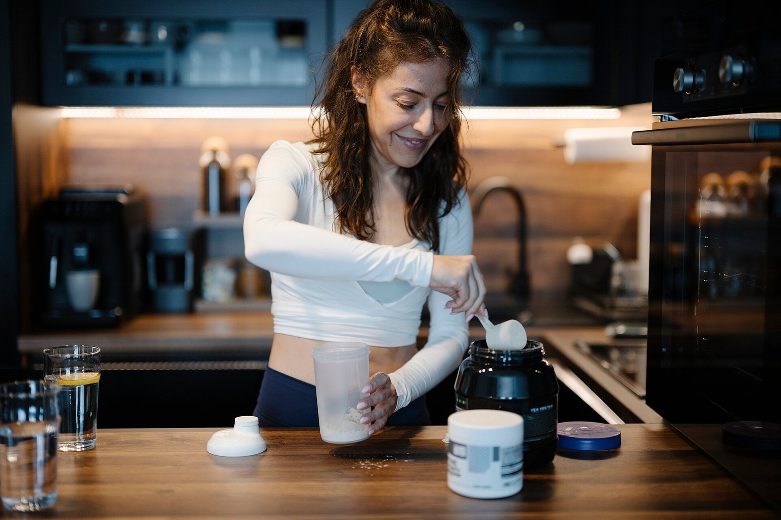 Woman scooping protein powder into a shaker bottle in a kitchen