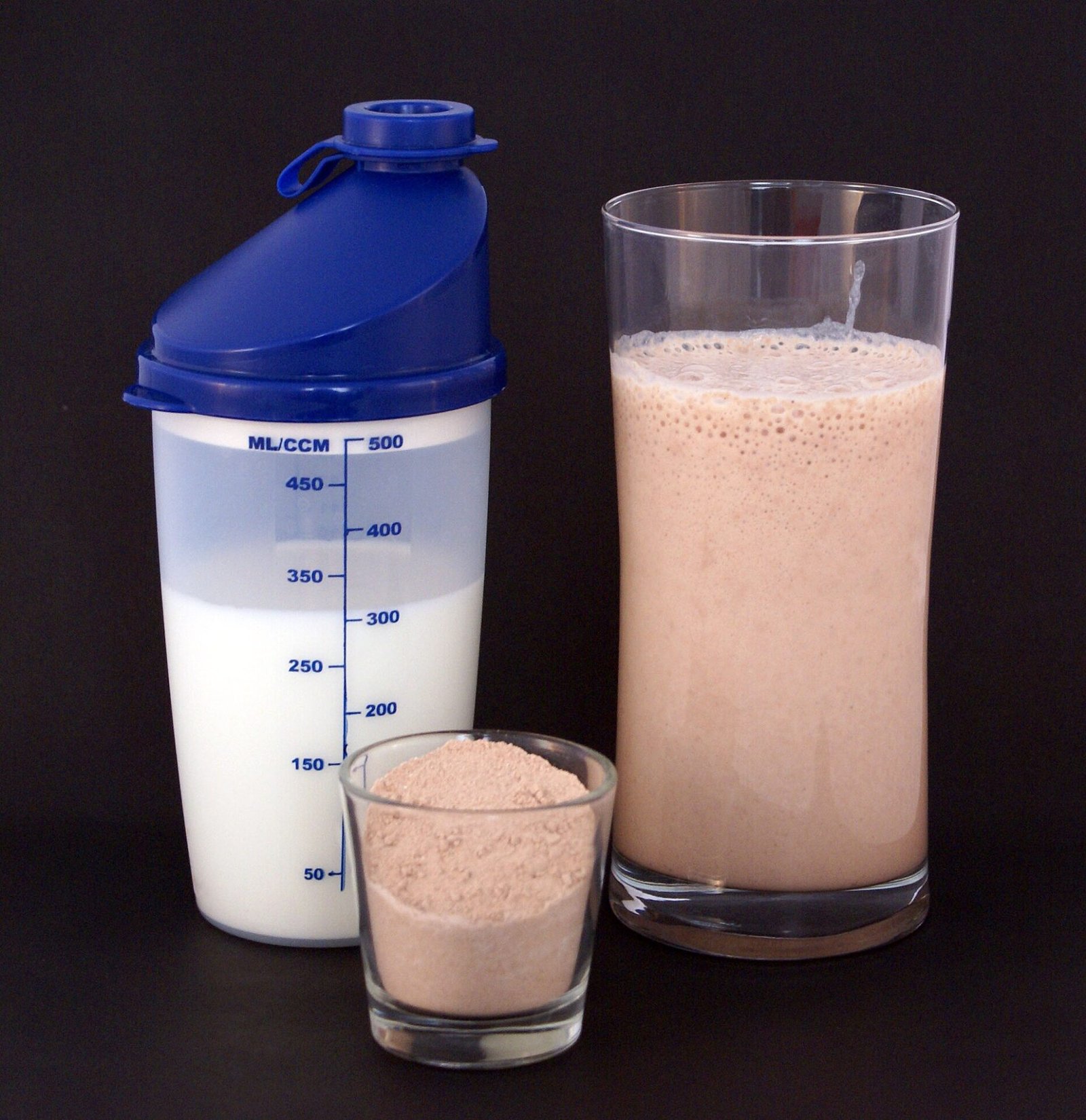 Protein powder, a shaker of milk and a finished protein shake on a dark background