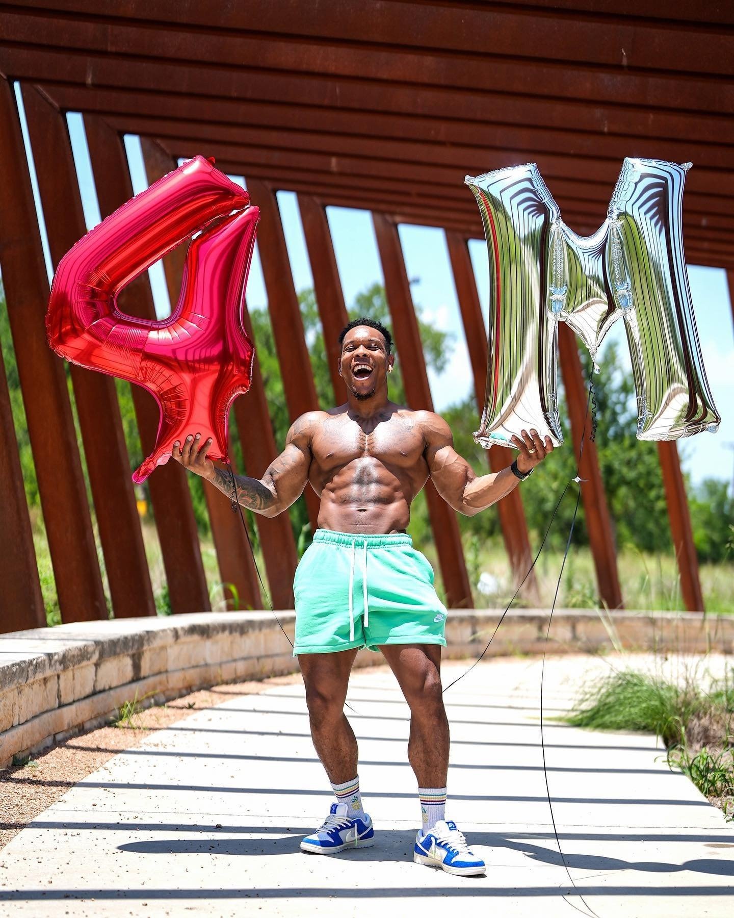 Bully Juice smiling shirtless outdoors holding red 4 and silver M balloons