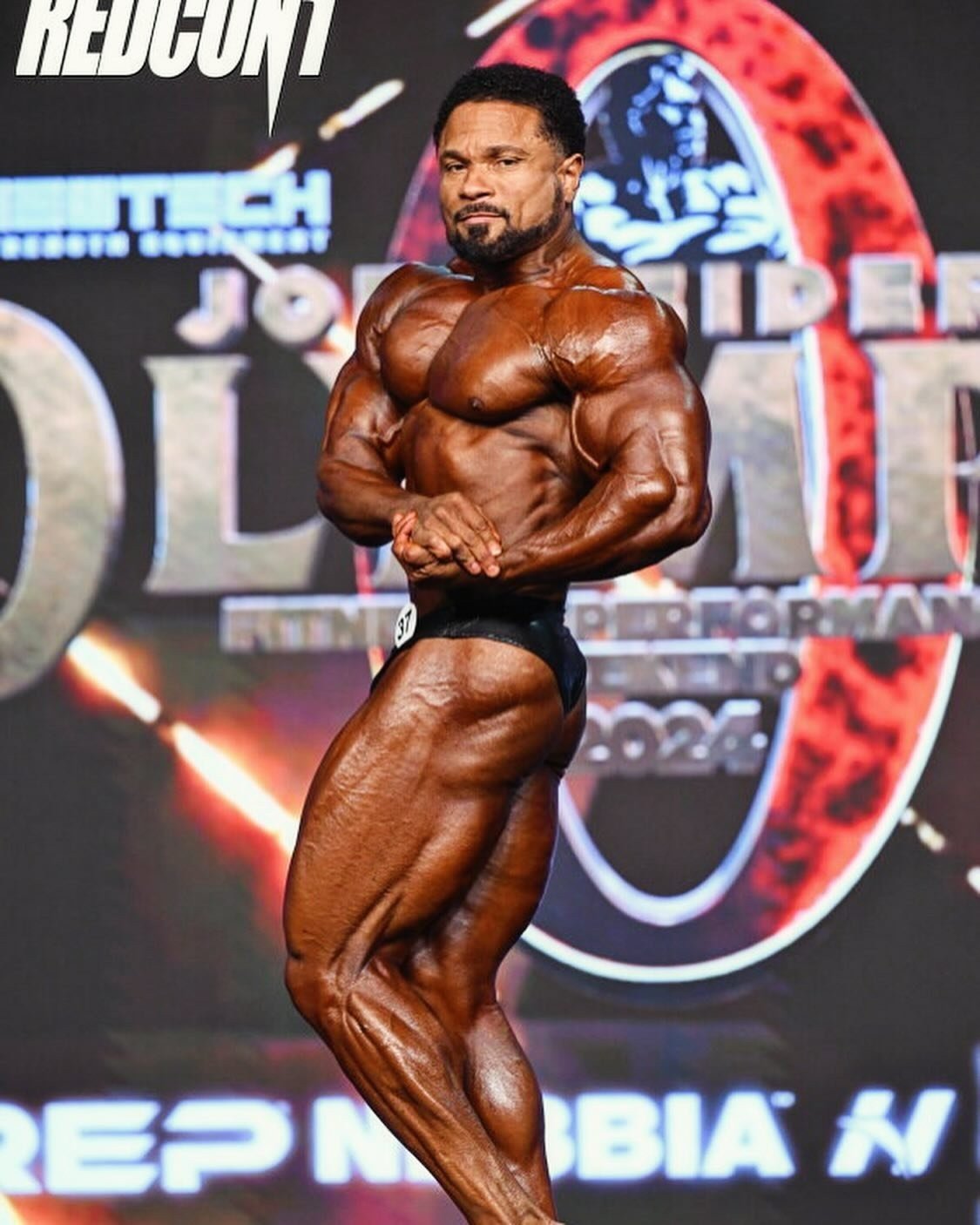 Damien Patrick performing a side chest pose on stage at the 2024 Mr. Olympia