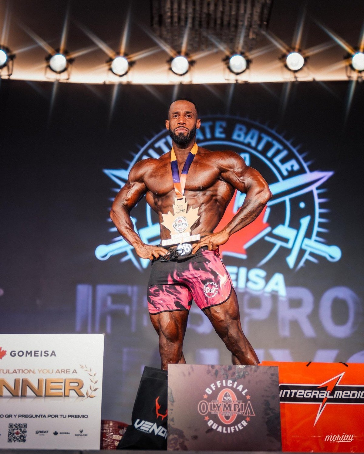 Deke Walker on the winner's podium at the 2025 Gomeisa Ultimate Battle Pro in Medellin, wearing his gold medal next to an Official Olympia Qualifier sign