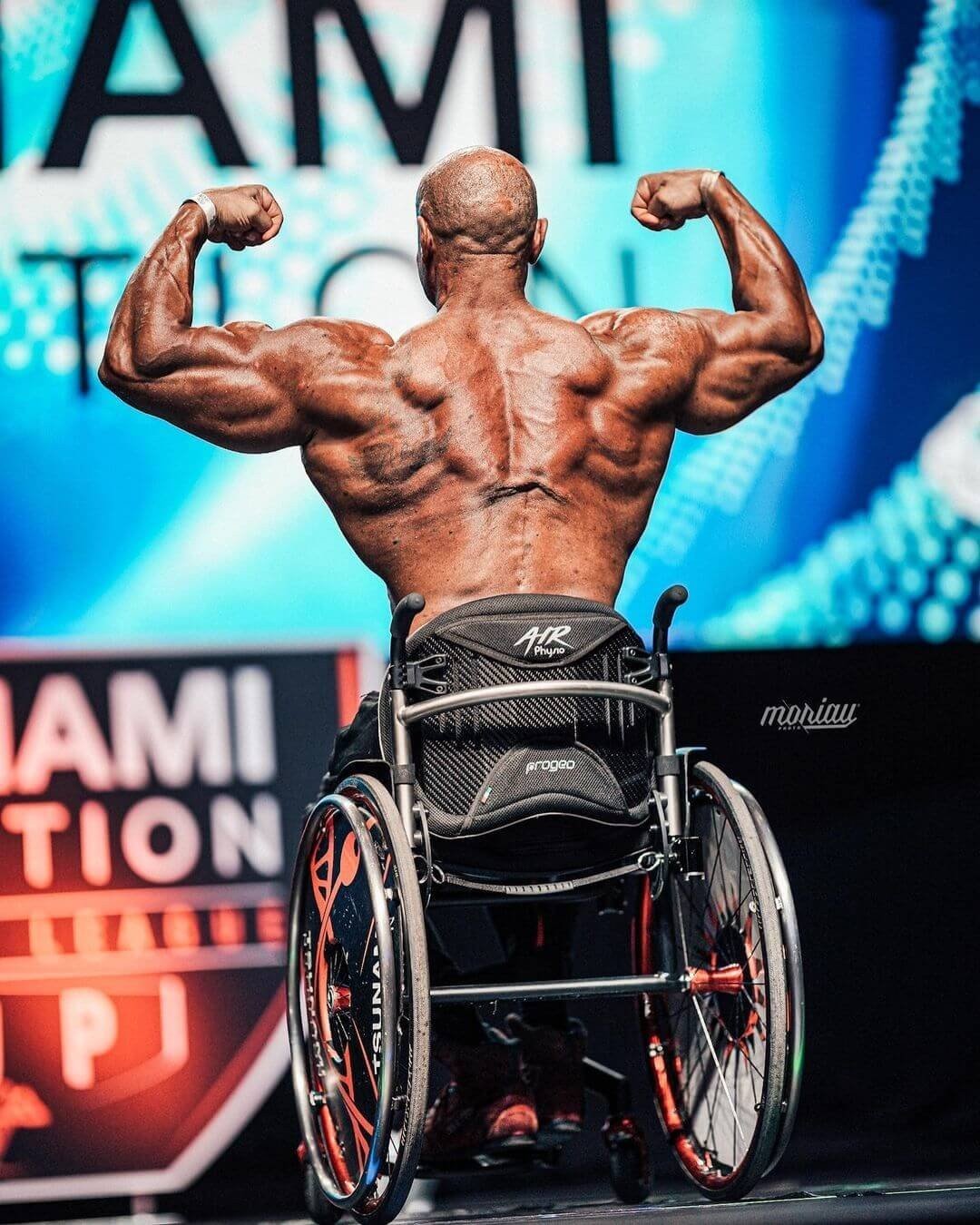 Gabriele Andriulli hitting a back double biceps pose on stage in his wheelchair