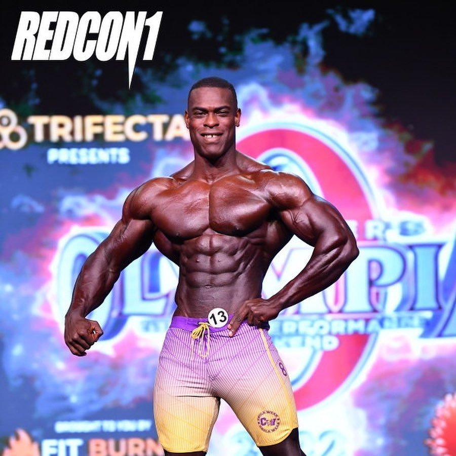Jeffrey Darko posing on the Mr. Olympia stage in purple and yellow board shorts
