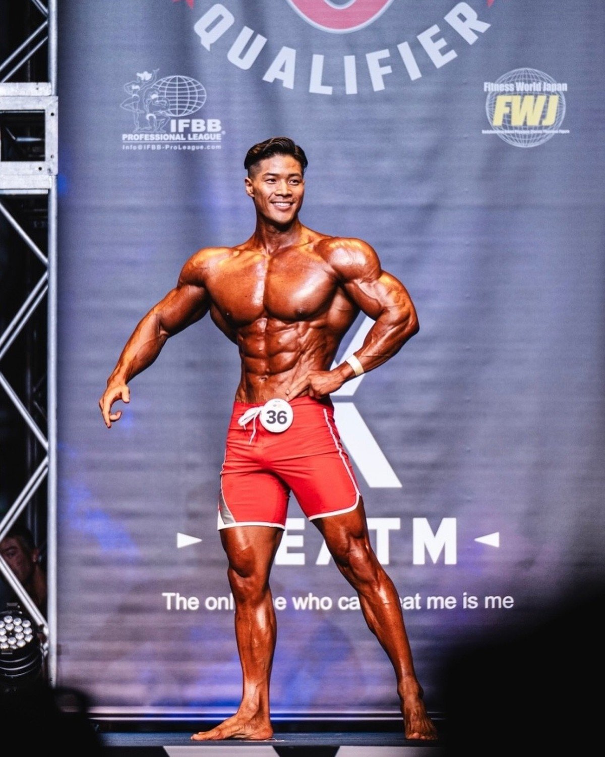Junpei Taguchi posing on stage in red board shorts at a Fitness World Japan IFBB Pro League event, competitor number 36