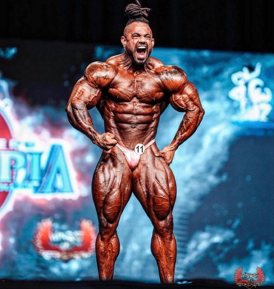 Lucas Coelho on stage, front lat spread, competitor number 11