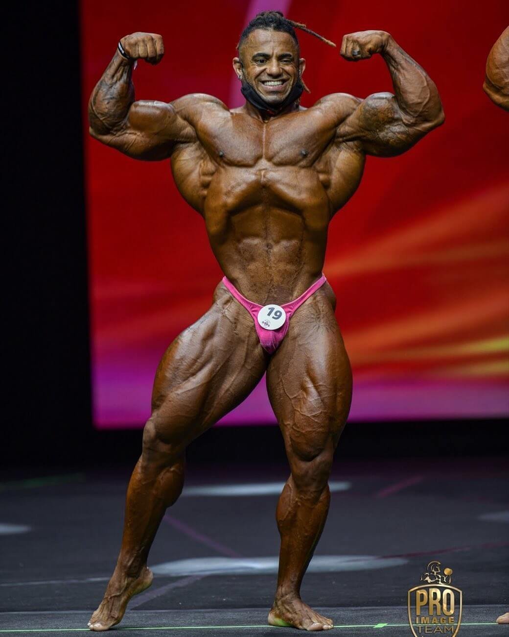 Lucas Coelho on stage, front double biceps, competitor number 19
