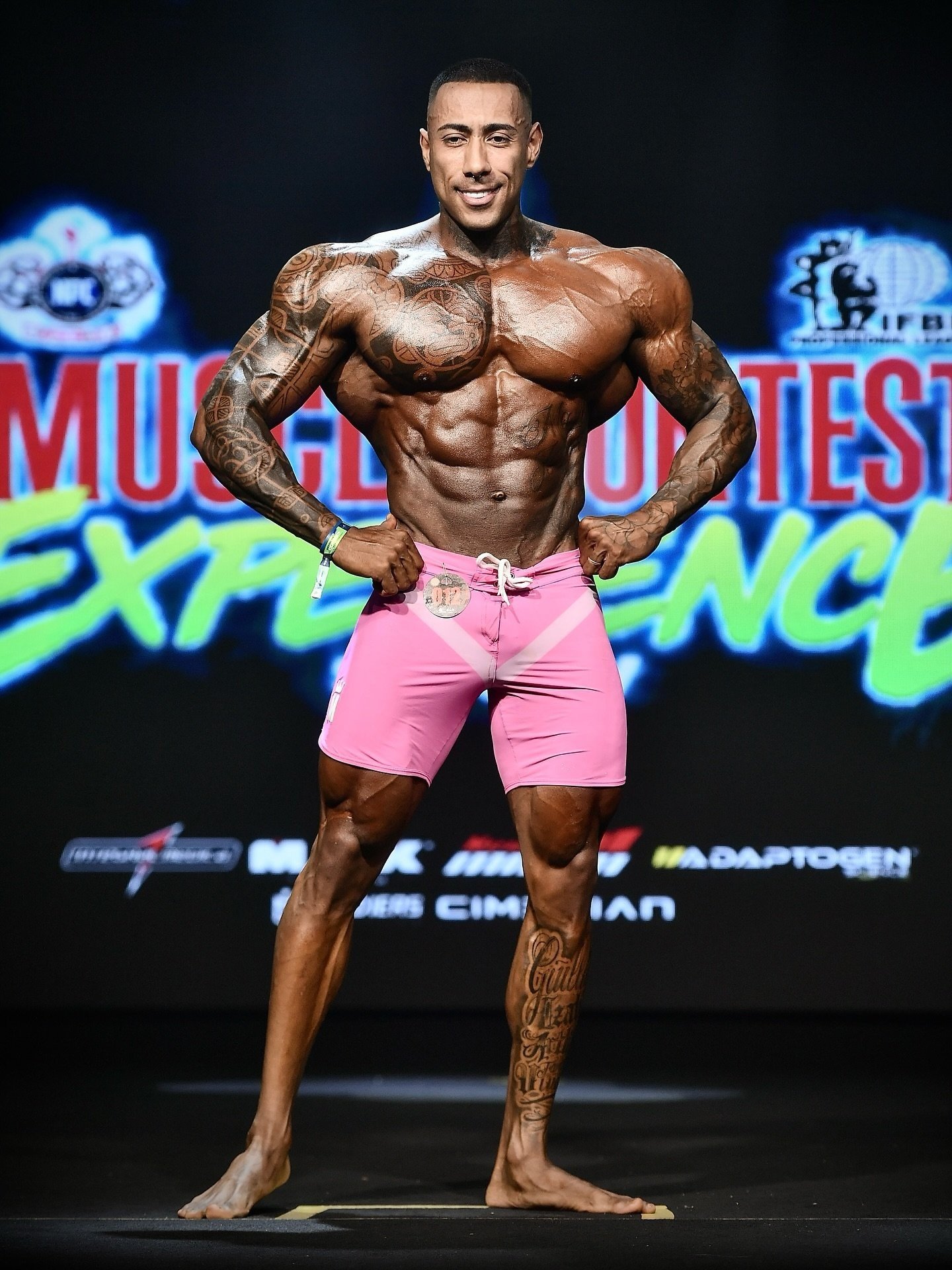 Lucas Viudes hitting a front pose on stage at the Musclecontest Experience Pro in pink board shorts