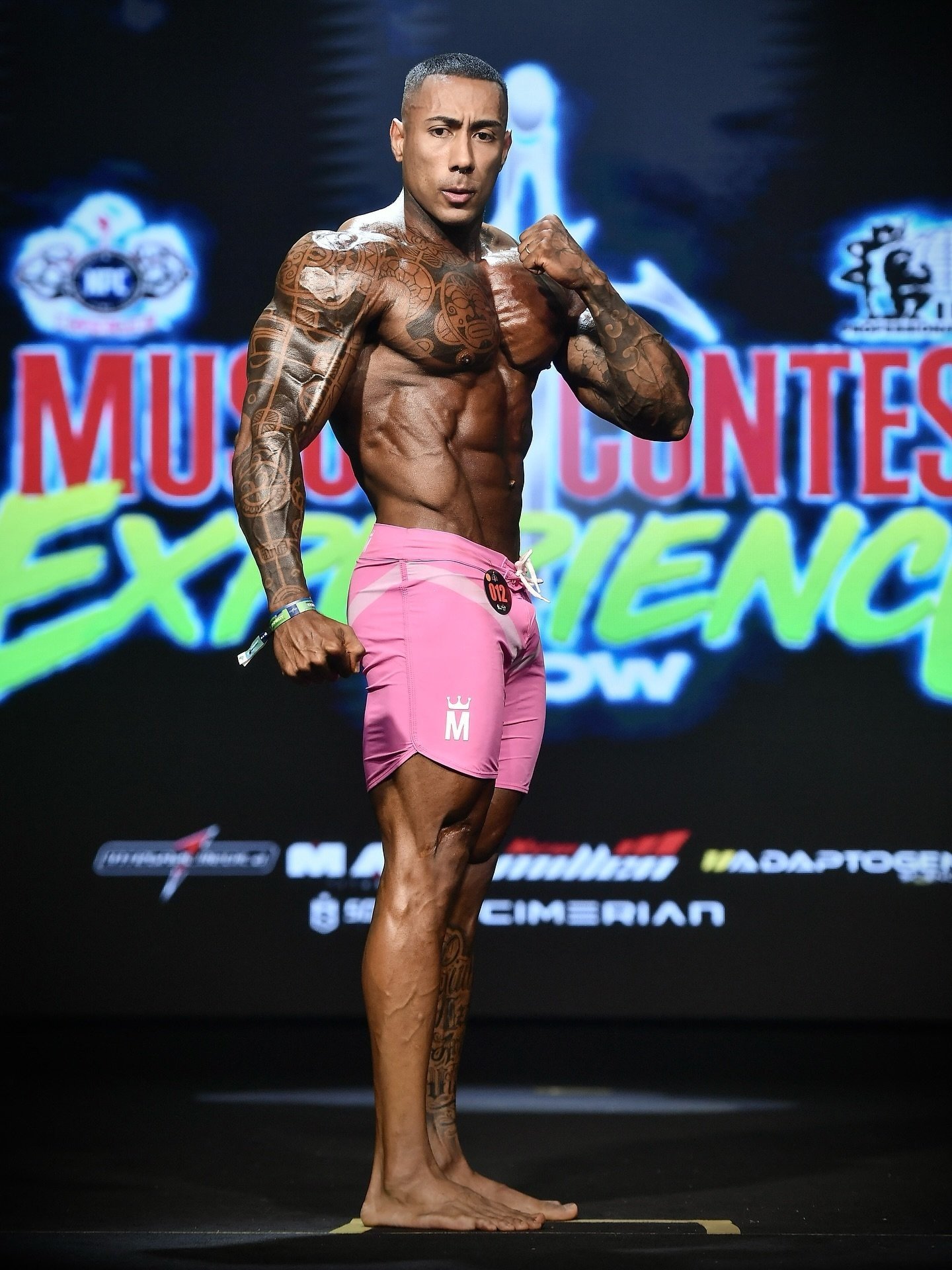 Lucas Viudes in a side pose on stage at the Musclecontest Experience Pro wearing pink board shorts