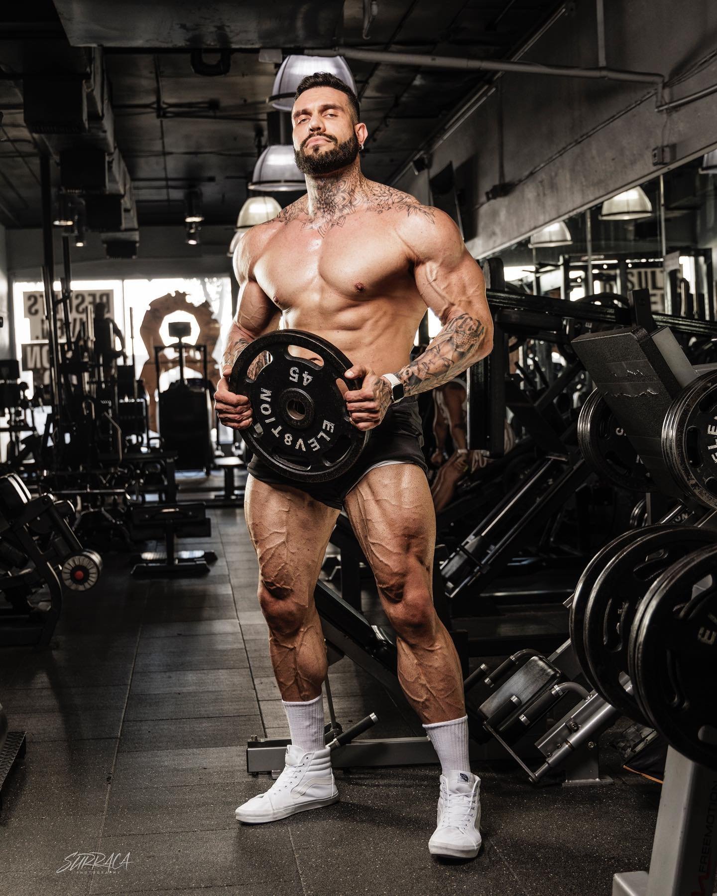 Lukas Lakutsin in the gym holding a 45 lb plate, showing his huge tattooed frame
