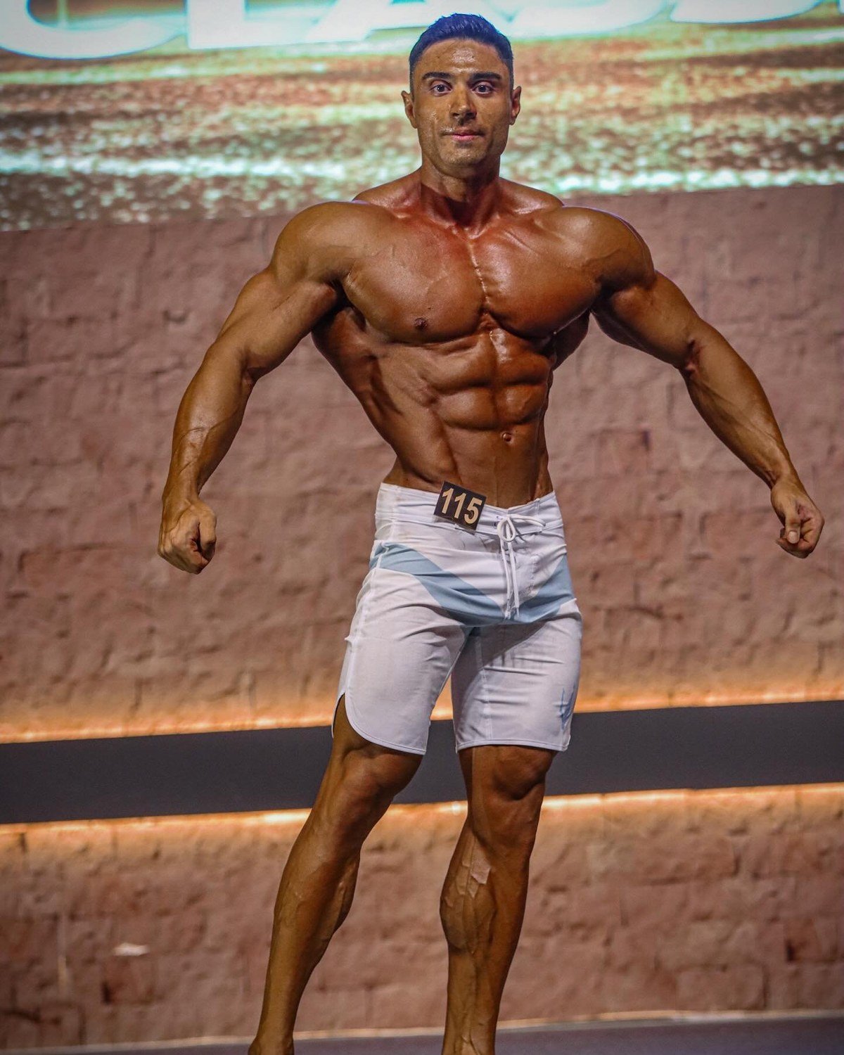 Ozcan Kirant hitting a front pose on the IFBB Pro League stage wearing competitor number 115
