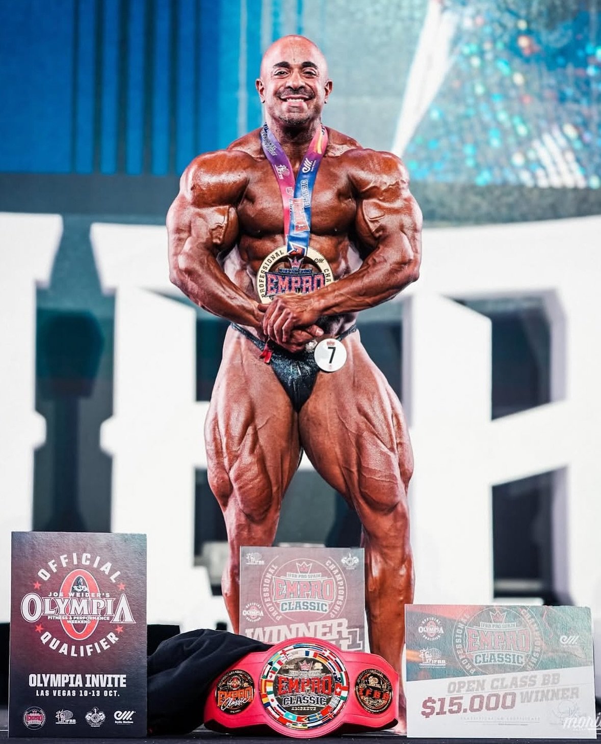 Sasan Heirati smiling on stage wearing his Empro Classic winner's medal and belt, next to his official Olympia qualifier invite